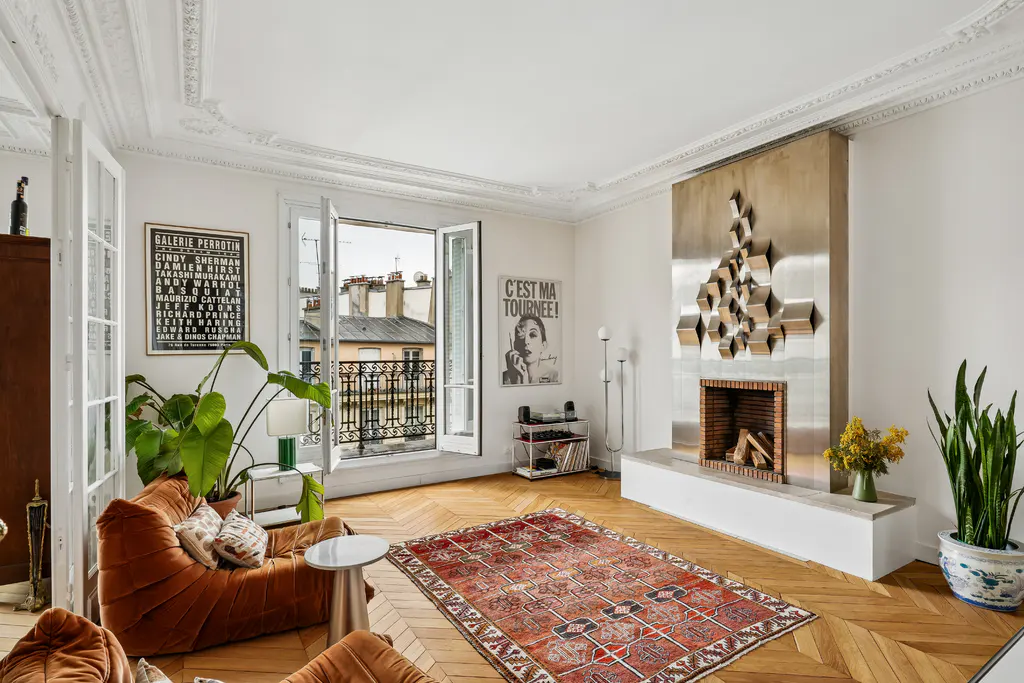 Bright living room with herringbone floors, a red patterned rug, and a modern fireplace with a geometric metal sculpture. Balcony doors open to a cityscape.