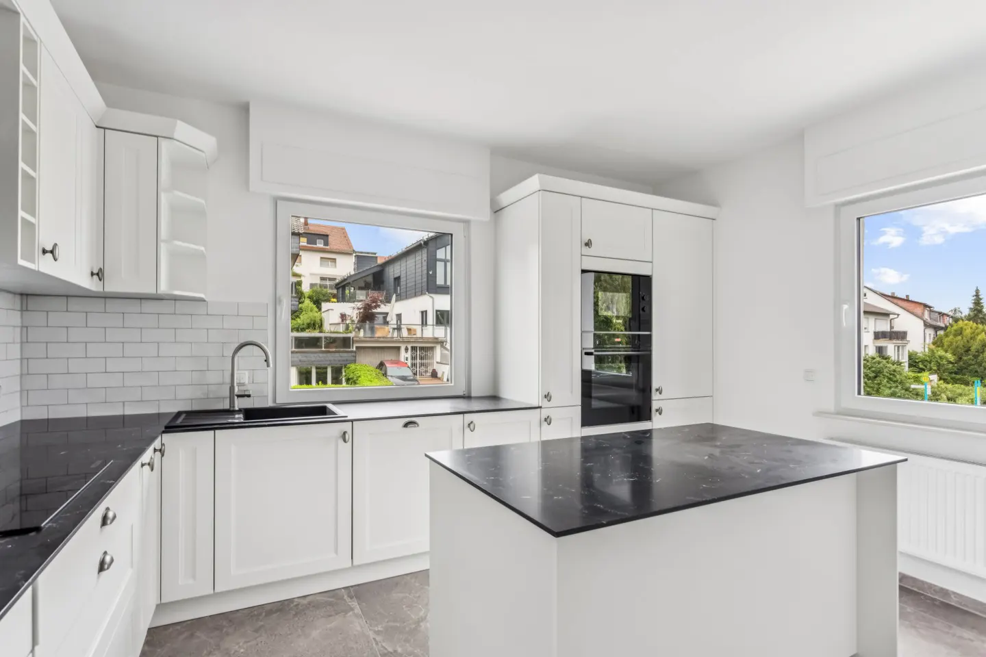Modern white kitchen with black marble countertops, a central island, subway tile backsplash, and views of houses outside.