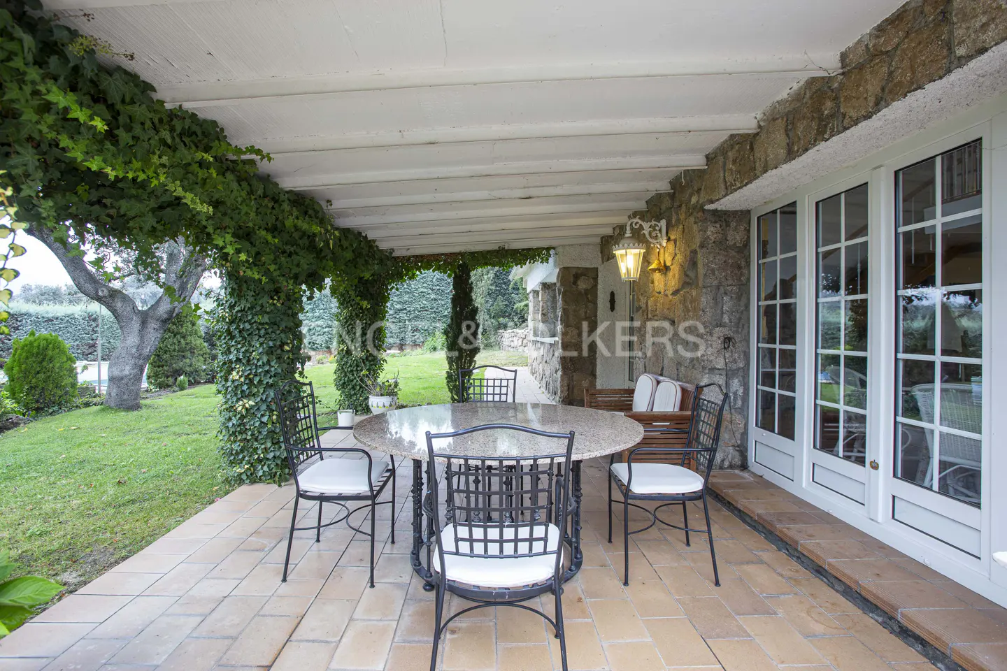Outdoor patio with stone walls, ivy-covered columns, and a round table with black chairs and white cushions.
