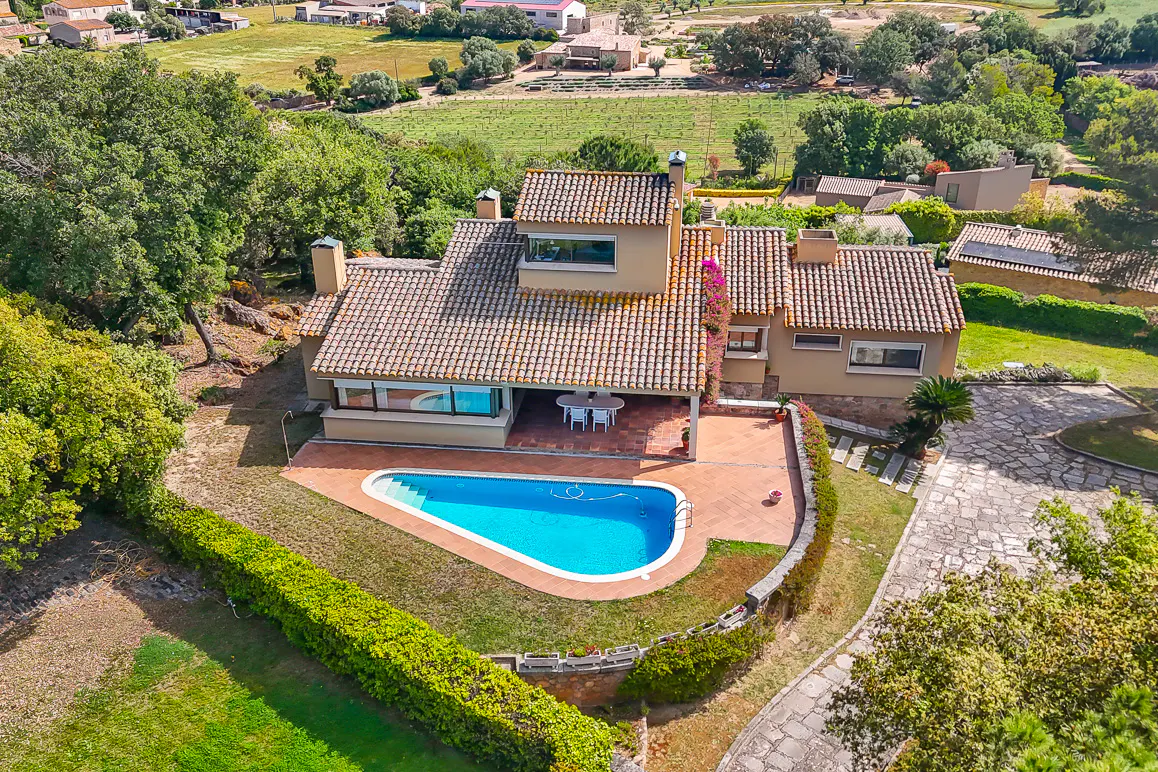 Aerial view of a tan house with a red tile roof, blue pool, and green landscaping.