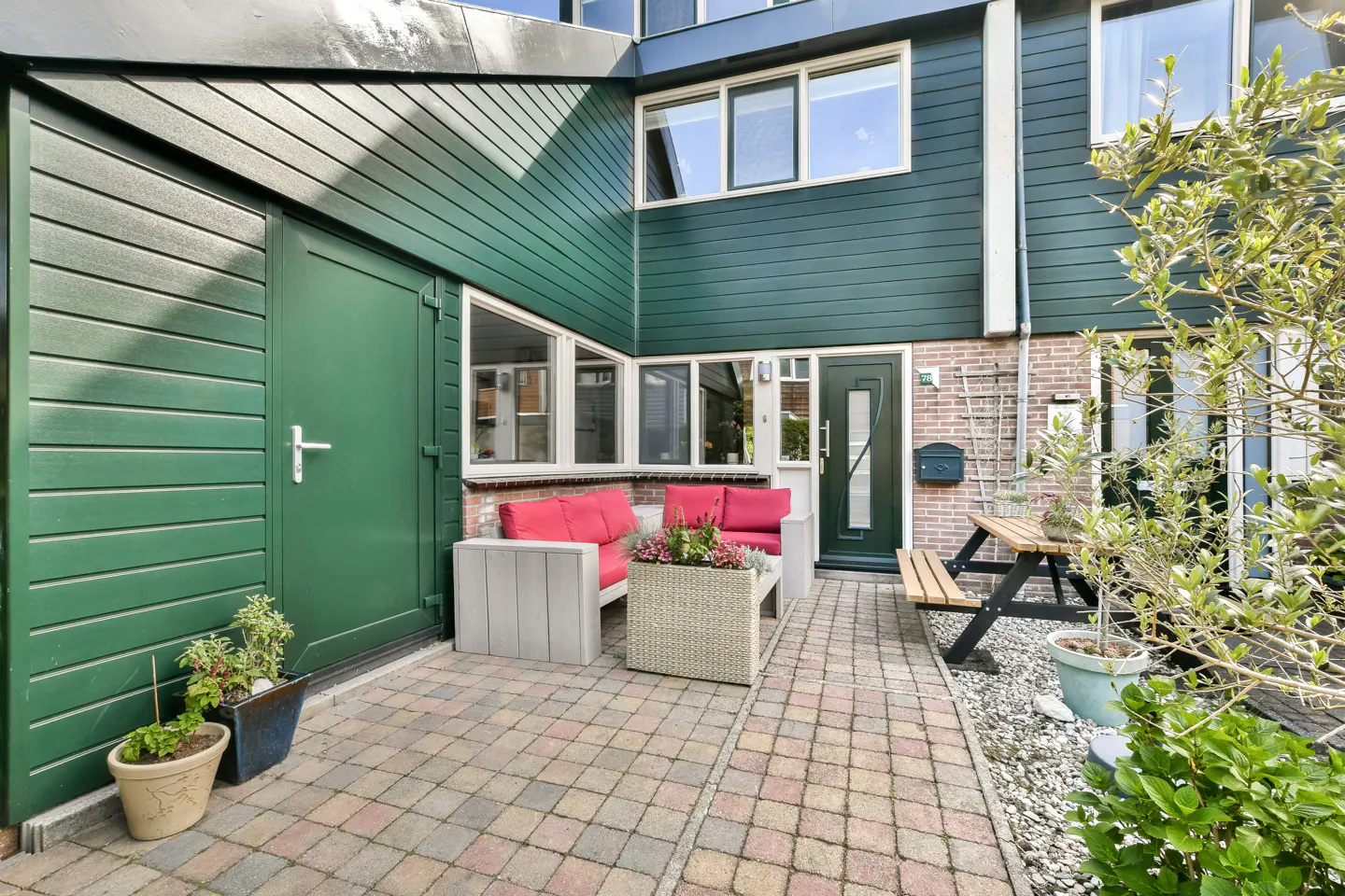 A green townhouse with a brick patio featuring outdoor seating, a picnic table, and potted plants.