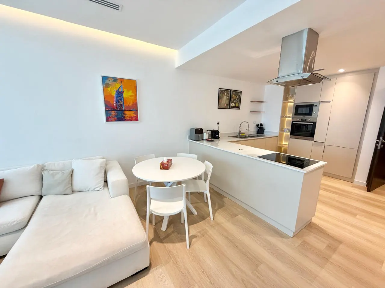 Bright, modern apartment interior with white walls, light wood floors, and a white sectional sofa. A round white table with chairs sits near an open kitchen with stainless steel appliances. An abstract painting adds color.