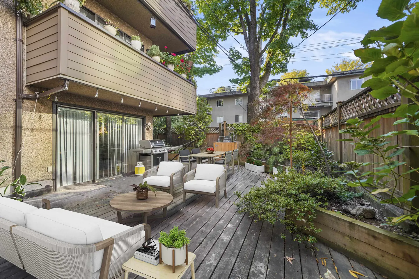A wooden deck with outdoor furniture, including a sofa, chairs, and a table, surrounded by lush greenery and a wooden fence.