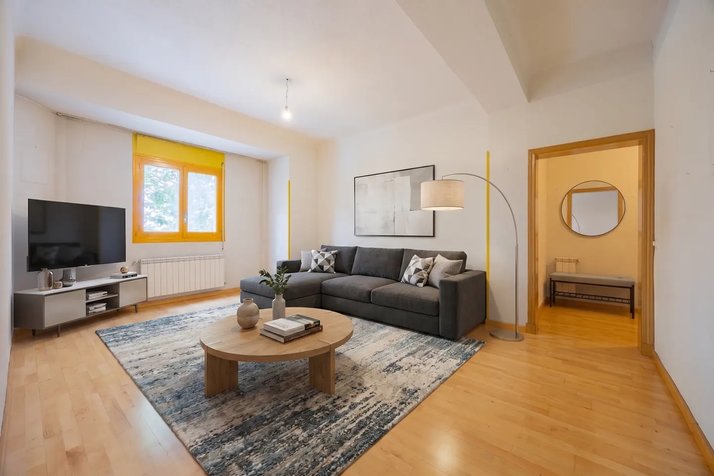 Bright living room with hardwood floors, a gray sectional sofa, and a round wooden coffee table on a blue patterned rug. A TV and yellow-framed window are visible.