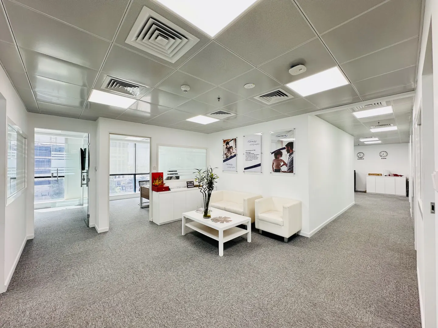 Bright office space with white walls, gray carpet, and a white reception area with seating. Posters adorn the walls.