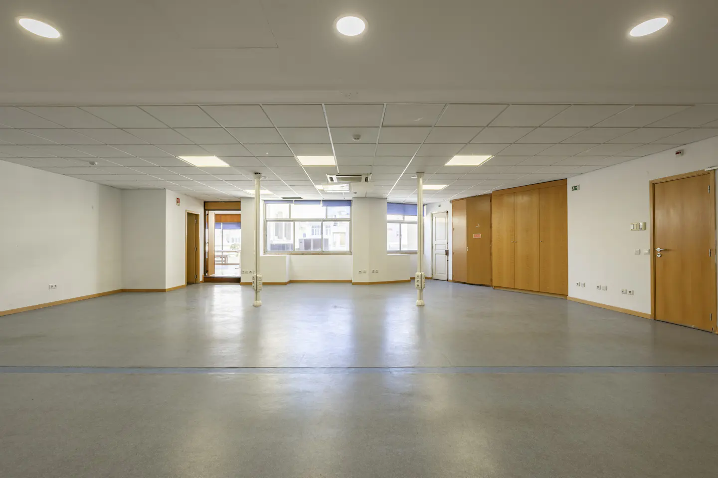 Empty room with gray floor, white walls, and a drop ceiling with square lights. Windows and wooden doors are visible.