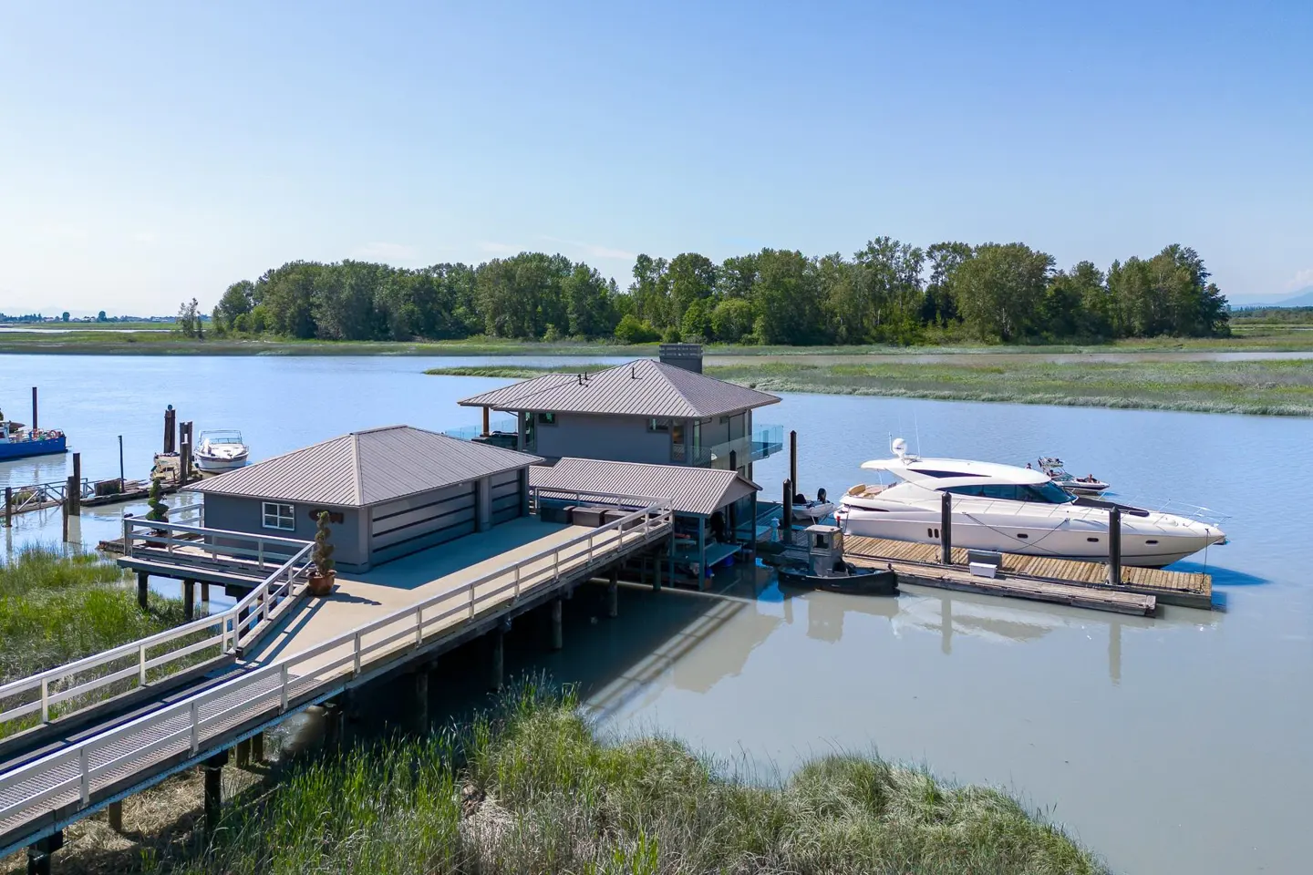 Waterfront home with a gray exterior and metal roof, connected by a wooden walkway to a dock with a yacht. Trees in background.