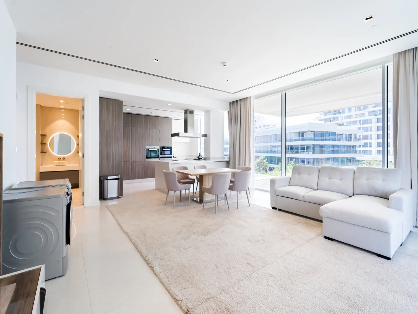 Modern open-plan living area with a white sectional sofa, dining table, and kitchen with wood cabinets and city views.