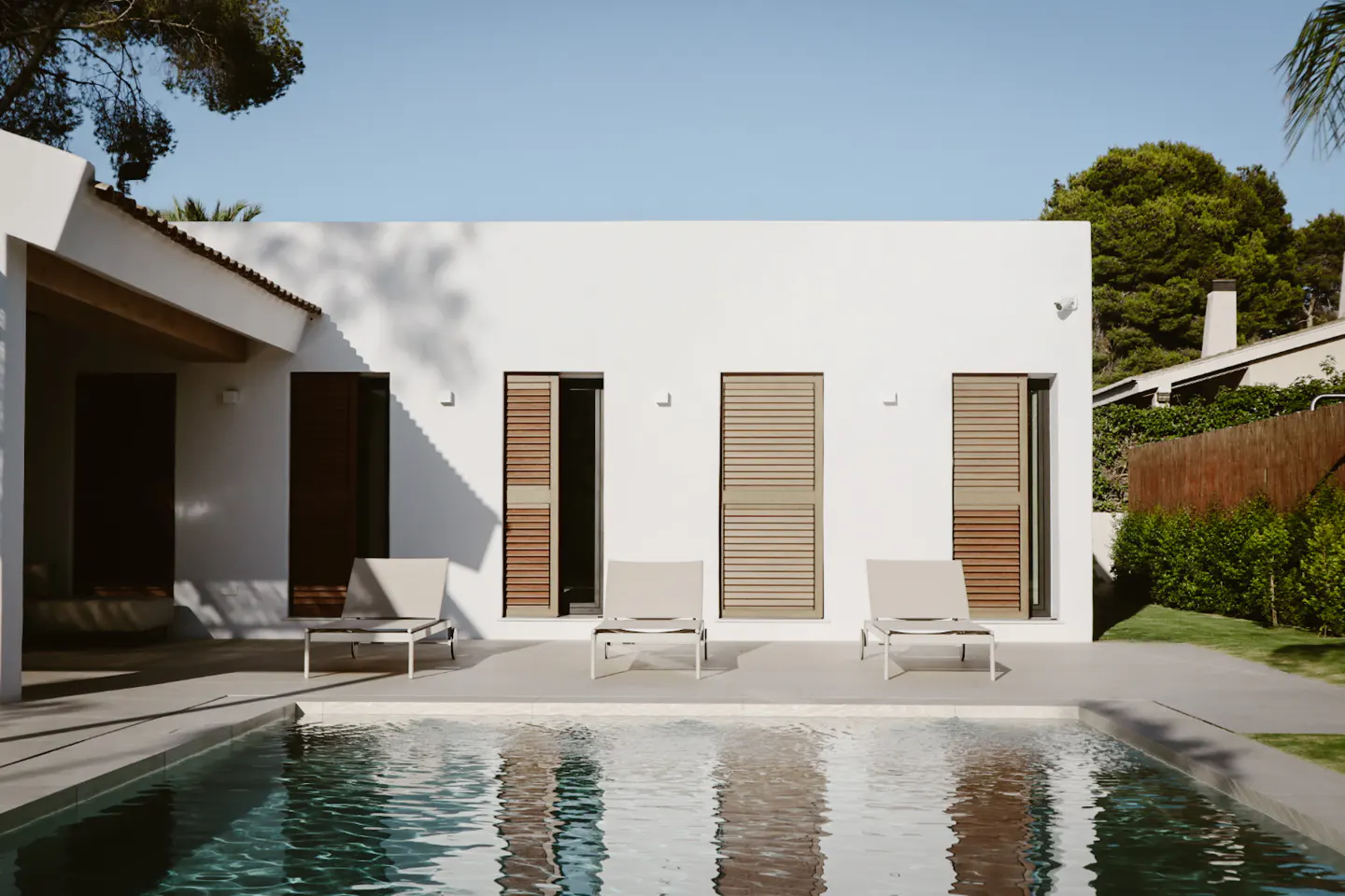 A modern white house with three lounge chairs by a pool. The windows have brown shutters.