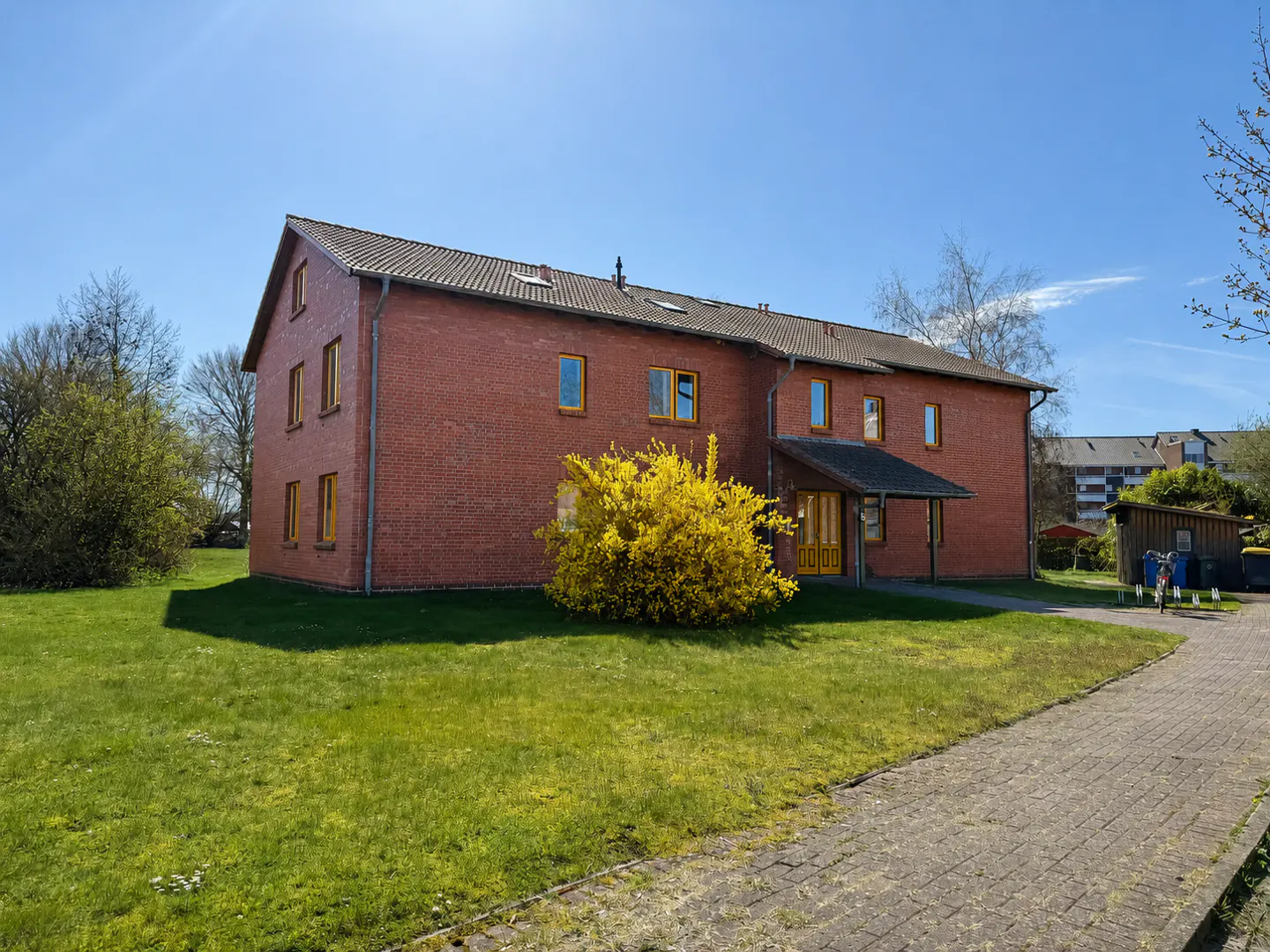 Two-story brick building with yellow window trim and a yellow forsythia bush in front, under a clear blue sky.