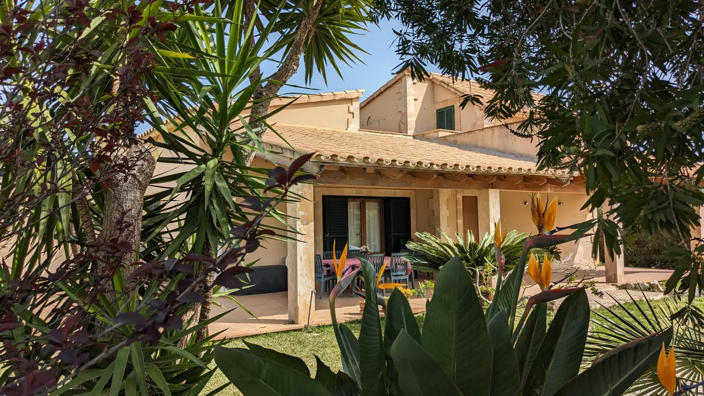 Exterior view of a tan house with a tiled roof, green shutters, and a covered patio with a table and chairs, framed by lush tropical plants.