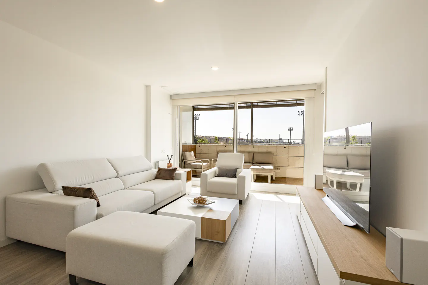 Bright living room with white walls, sofa, chair, and a balcony with city views. A modern TV stands on a wooden cabinet.
