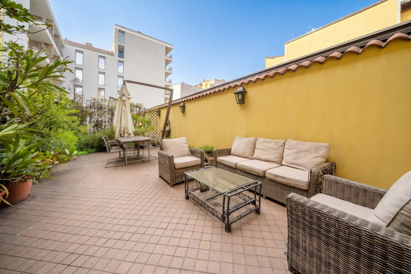 Outdoor patio with wicker furniture, including a sofa, chairs, and a glass-topped coffee table, against a yellow wall.