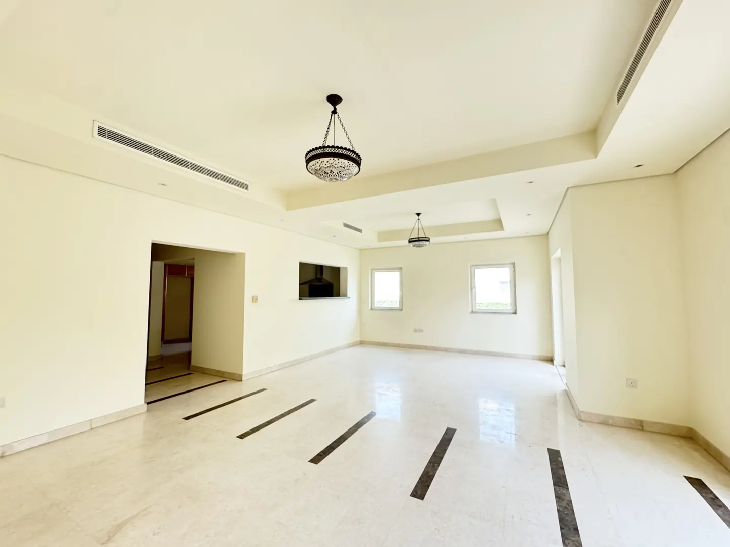 A bright, empty living room with marble floors and two ornate chandeliers. Two windows and a kitchen pass-through are visible.