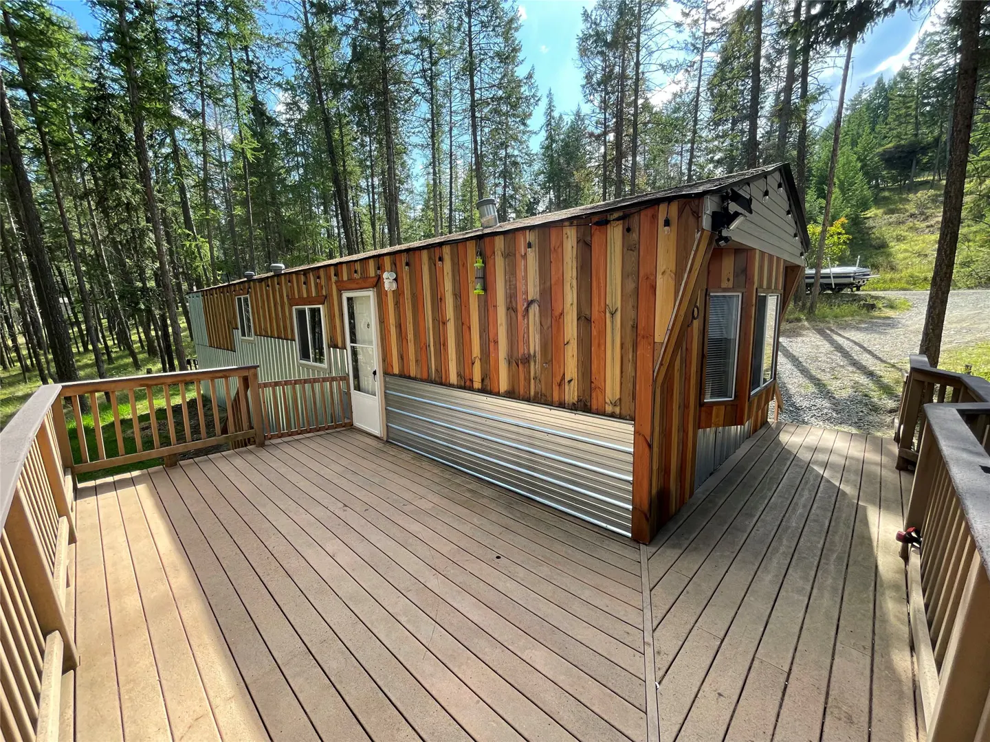 Exterior view of a wood cabin with a deck, surrounded by tall trees. The cabin has a mix of wood and metal siding.