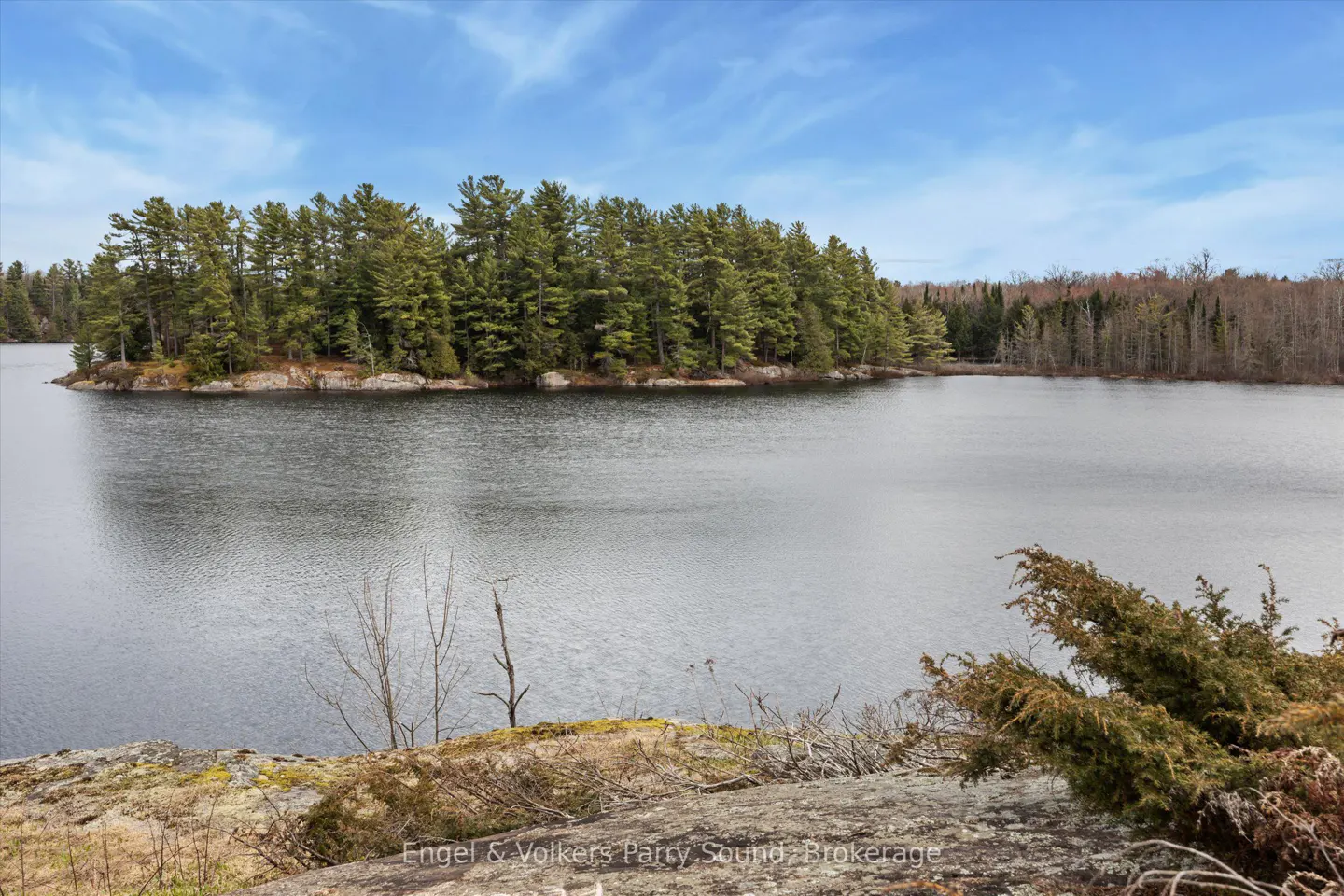 Scenic view of a calm lake with a tree-covered island under a blue sky with wispy clouds. Foreground shows rocky terrain and greenery.