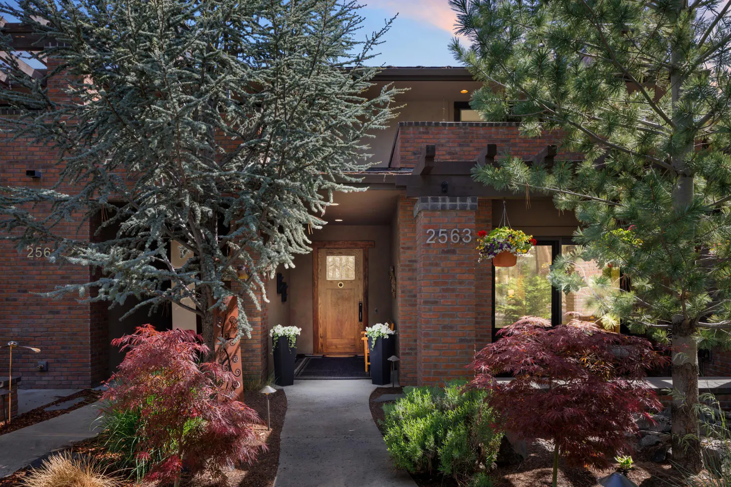 A brick home entrance with a wooden door, concrete walkway, red Japanese maples, and potted white flowers.