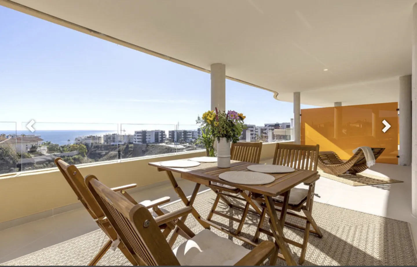 Balcony with wood table, chairs, and flowers overlooking the ocean and city. A lounge chair sits in the background.