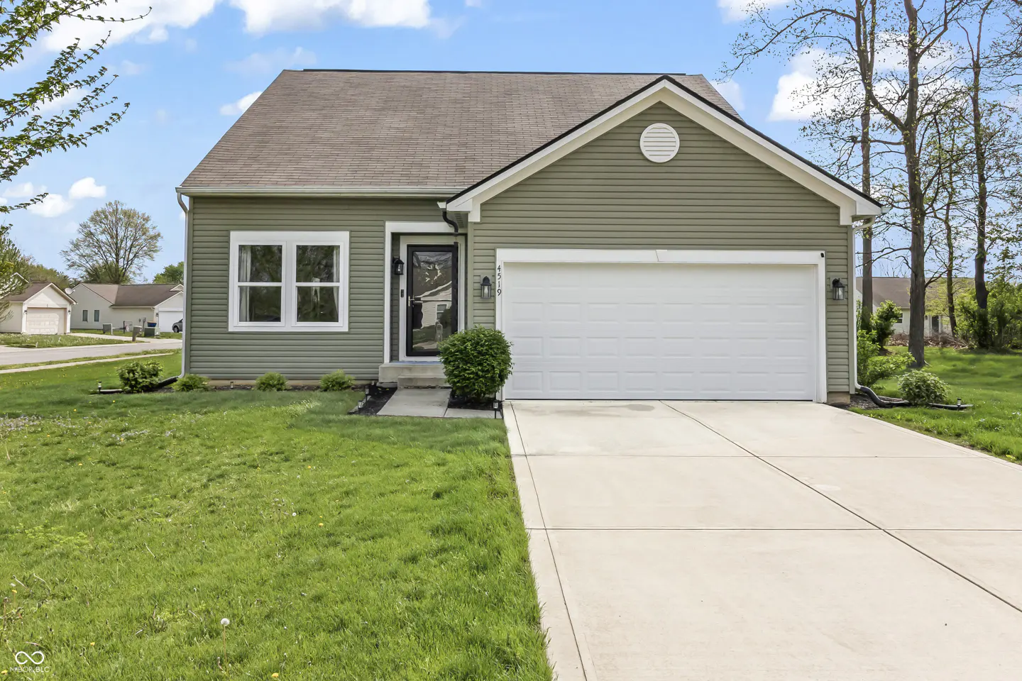 A single-story home with green siding, a brown roof, and a white garage door sits on a green lawn under a blue sky.