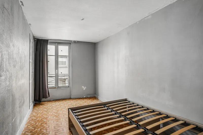 Empty gray bedroom with a wooden bed frame, patterned floor, and window with gray curtains.