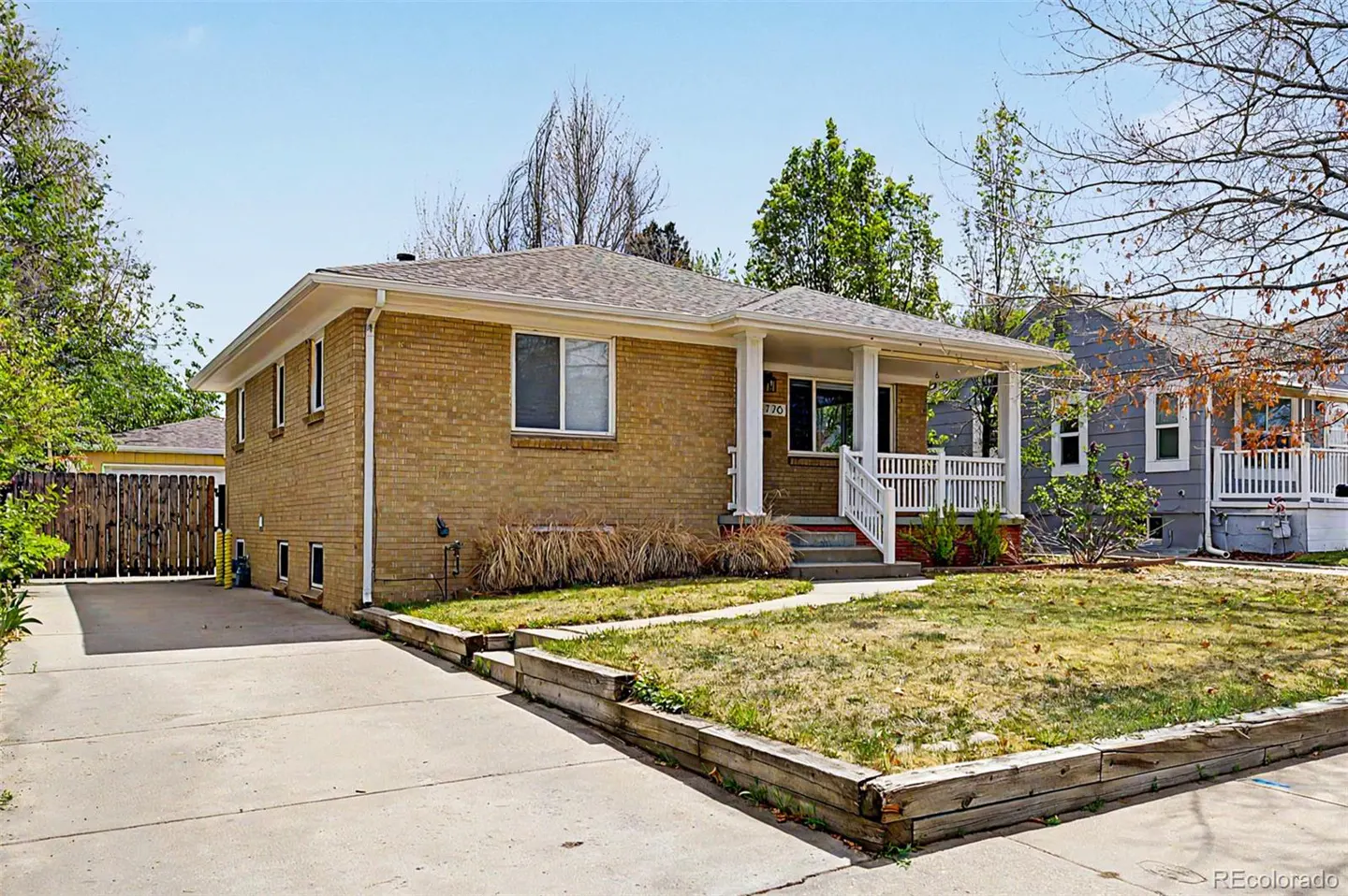 A tan brick house with a white porch and a concrete driveway on a sunny day.