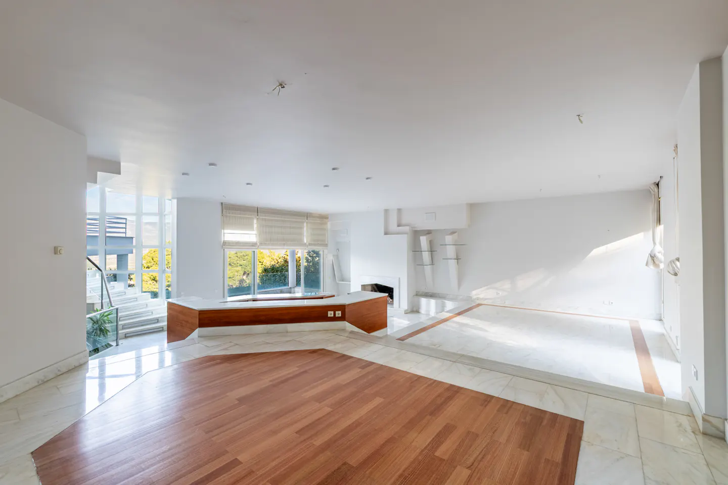 Bright, empty living room with wood and marble floors, white walls, and large windows with blinds.