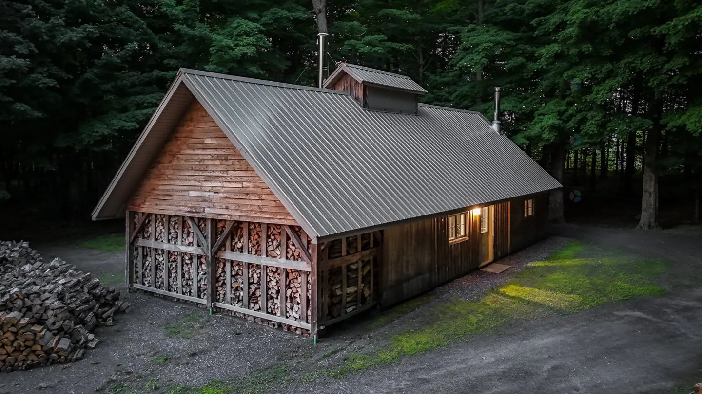 Rustic wood cabin with a metal roof, surrounded by trees. Wood is stacked under the front porch. Light shines from the windows.