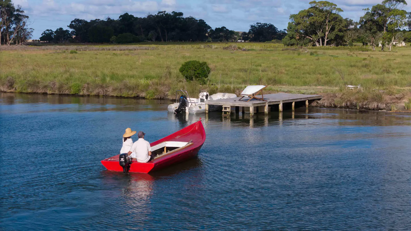 A red boat with two people on a blue river, with a dock and green fields in the background.