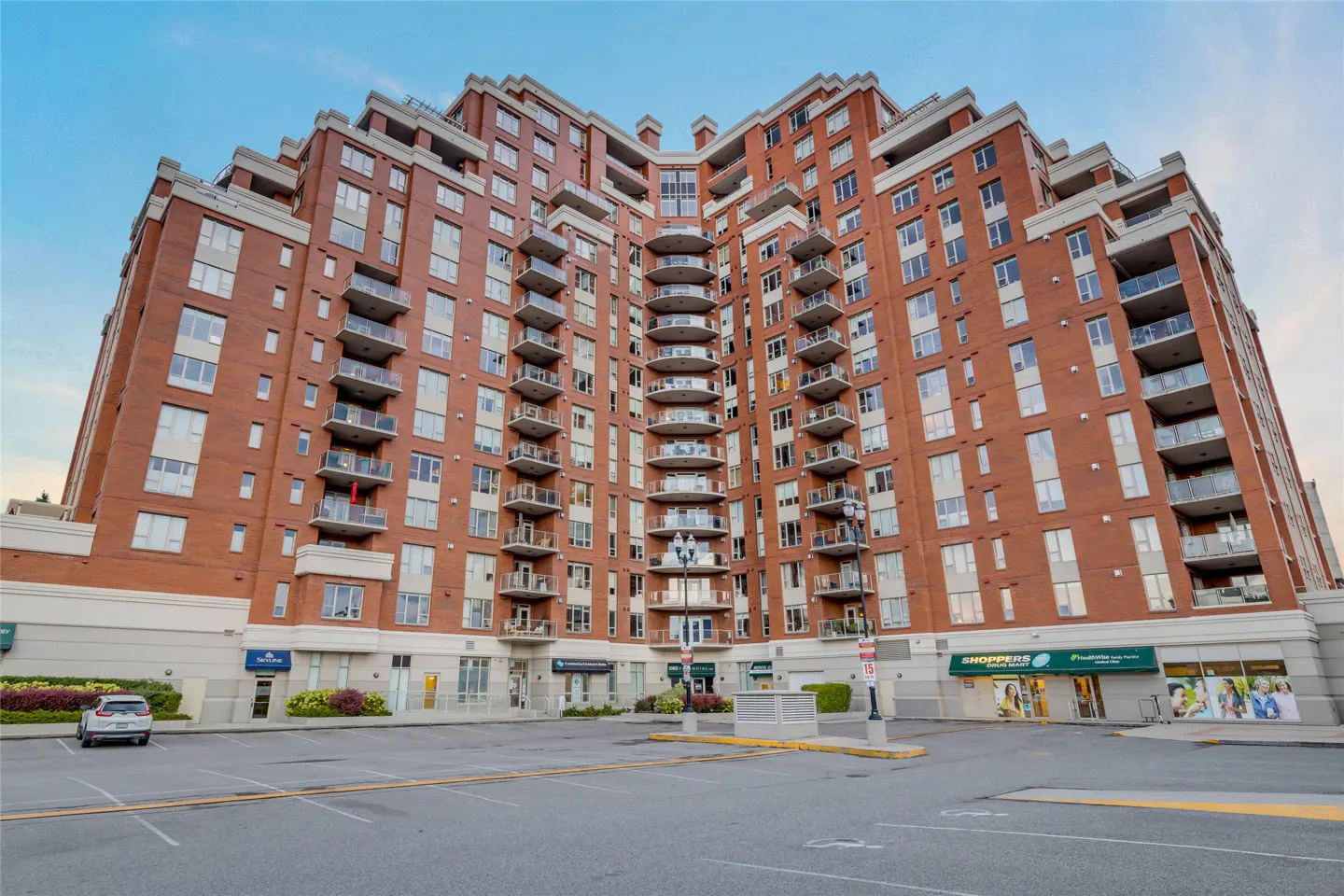 Exterior view of a tall, red brick apartment building with balconies and a parking lot in front. Shoppers Drug Mart is visible.