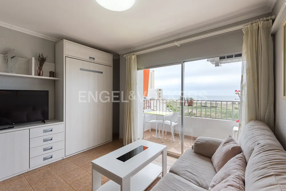 Living room with a beige sofa, white coffee table, and a balcony overlooking the ocean. A TV and built-in storage are on the left.