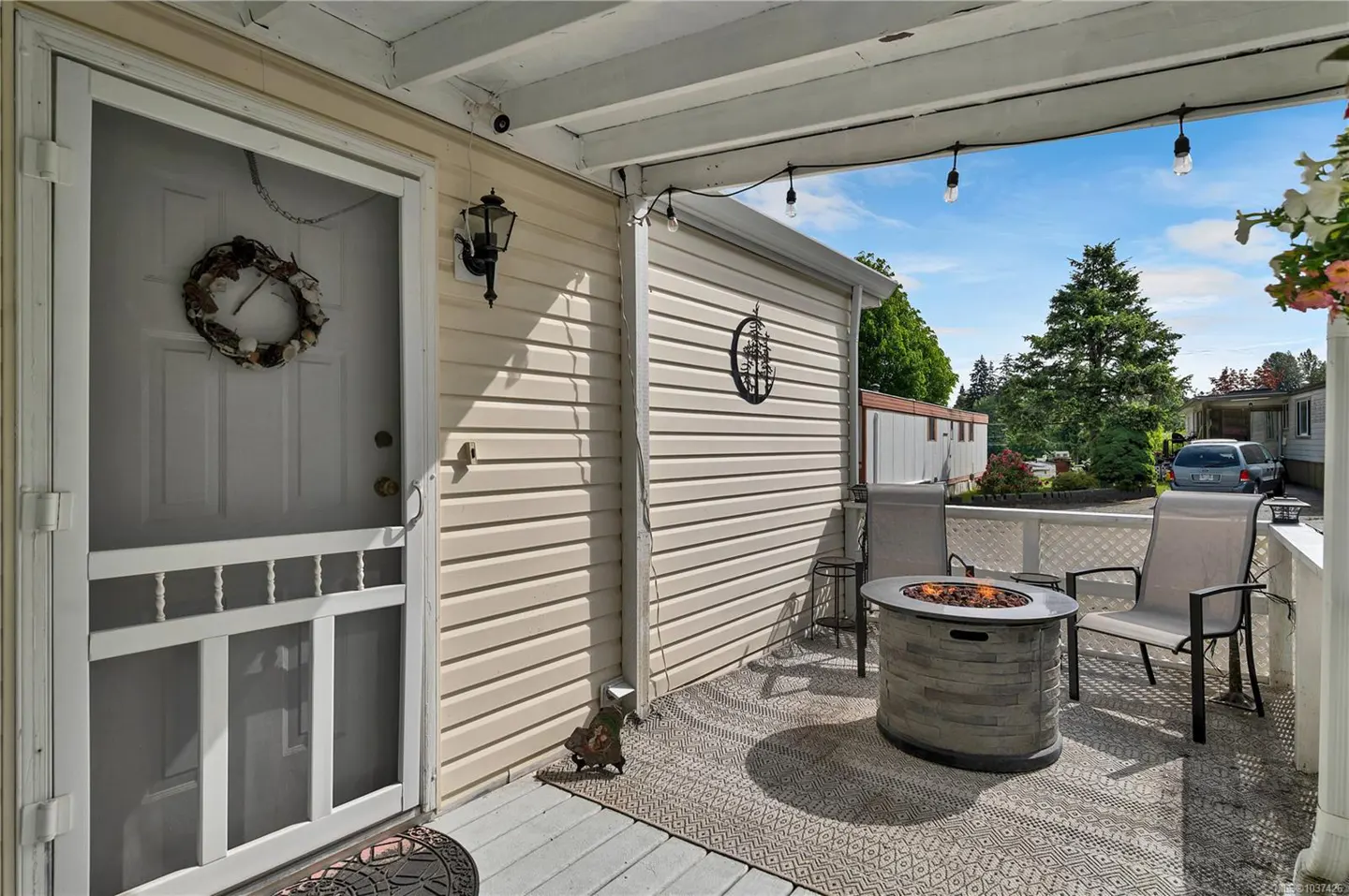 Covered porch with a white door, wreath, and a fire pit with chairs. String lights hang above, with a view of trees and a car in the background.