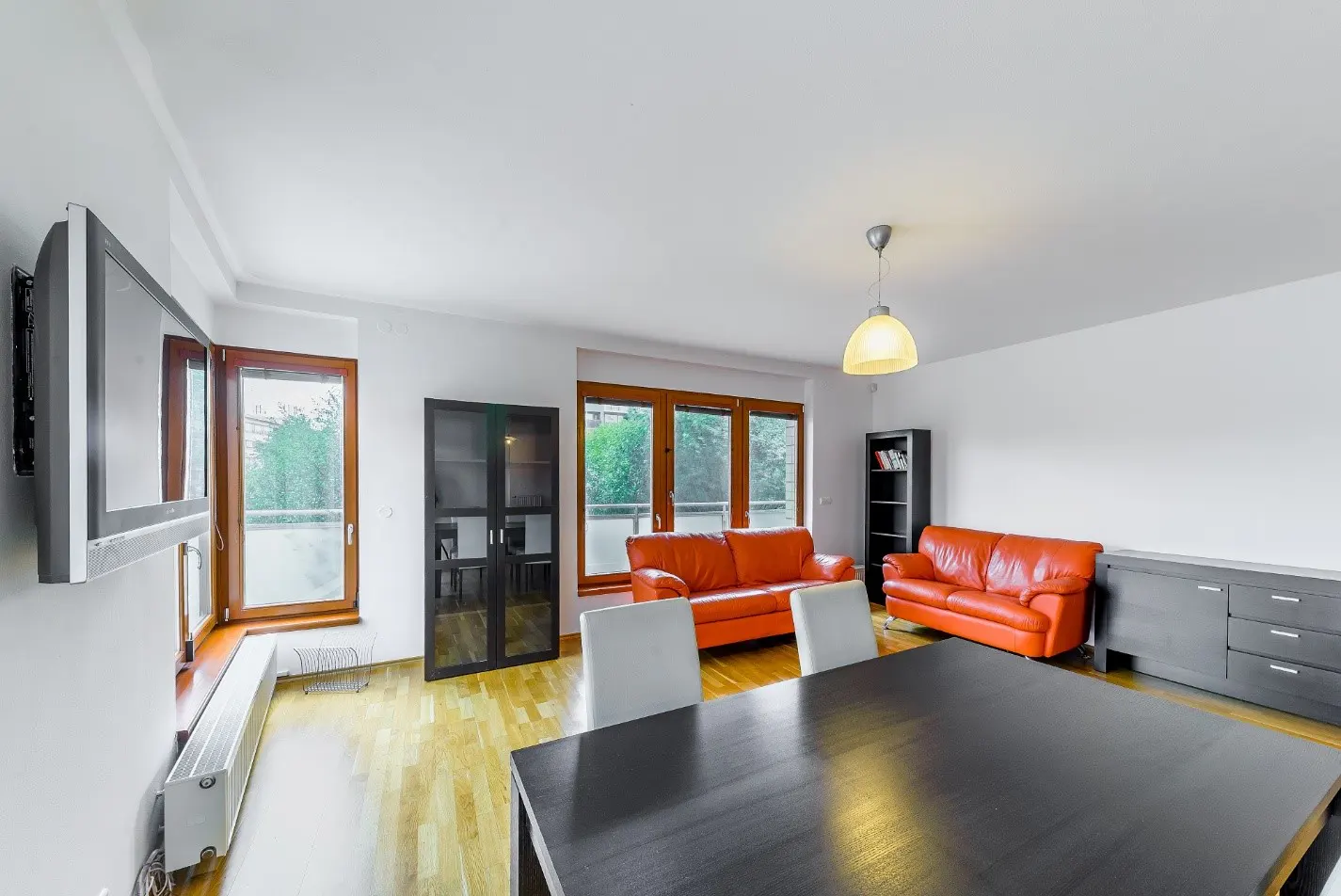 Bright living room with hardwood floors, orange sofas, a dark wood dining table, and large windows.