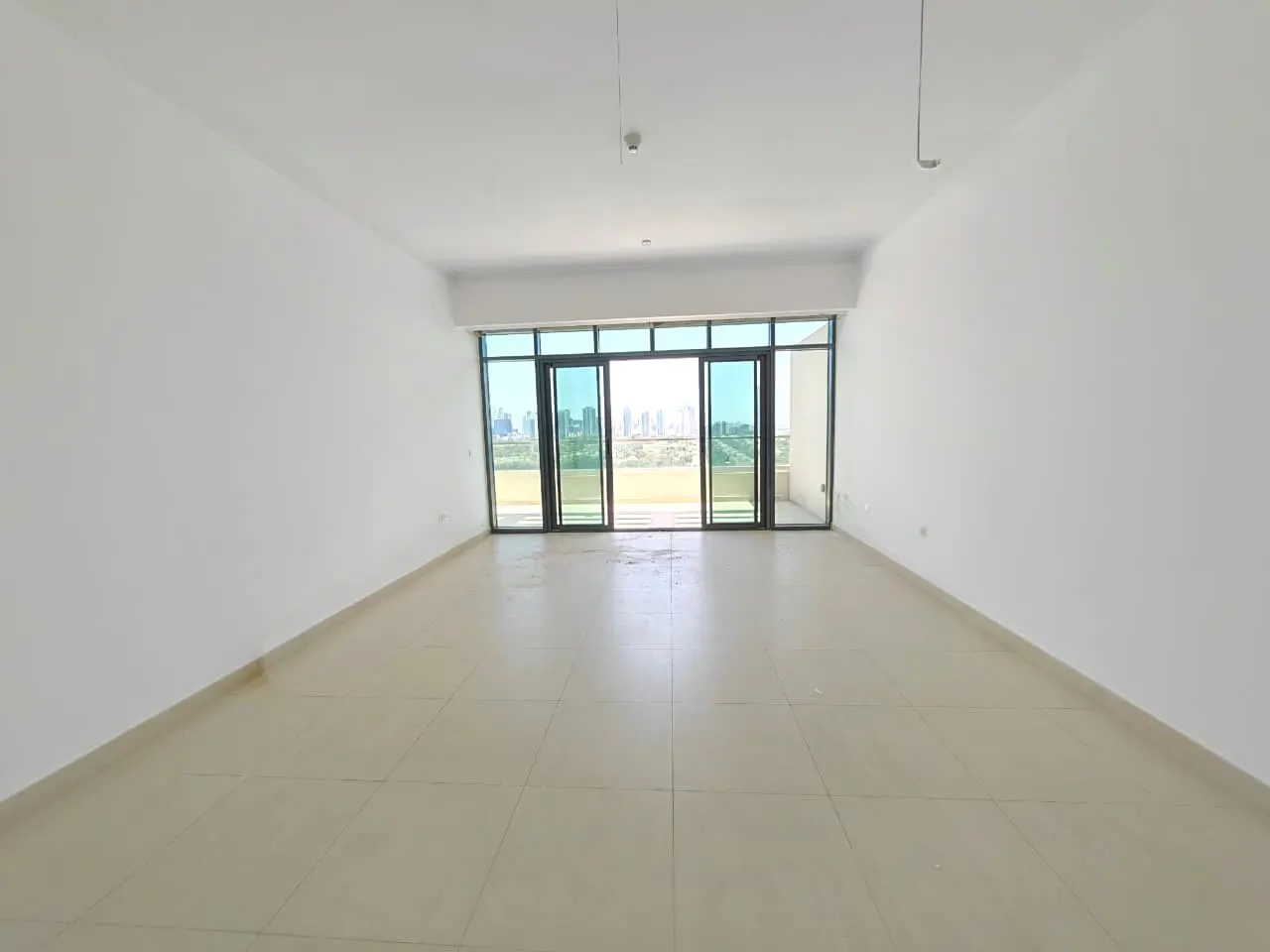 Empty room with white walls and beige tile floor. Sliding glass doors open to a balcony with a city view.