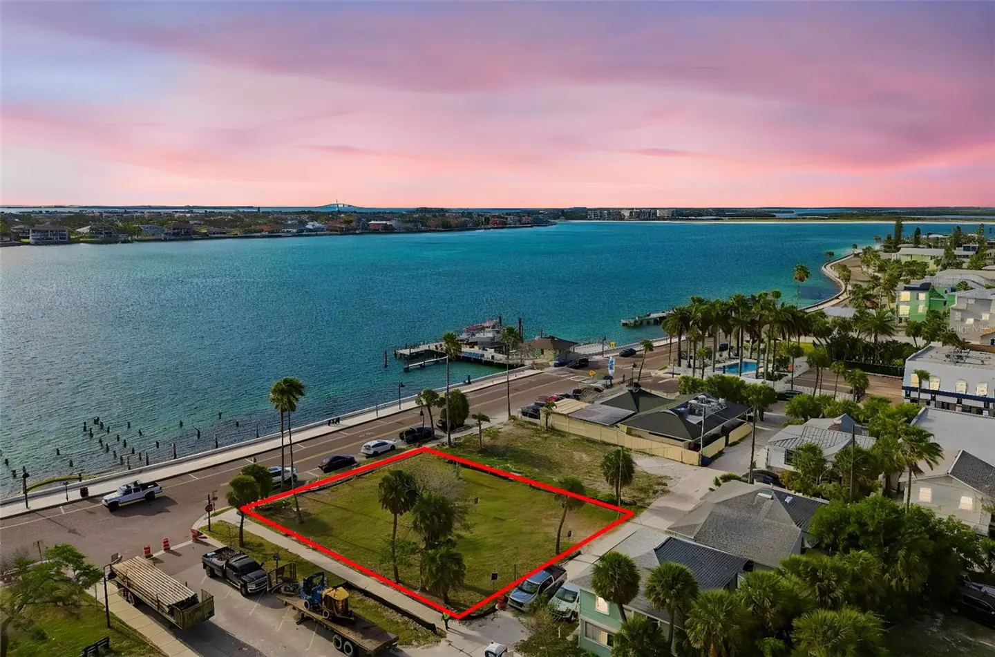 Aerial view of a grassy lot outlined in red, near a waterfront road with palm trees, cars, and a pier, under a pink and purple sky.