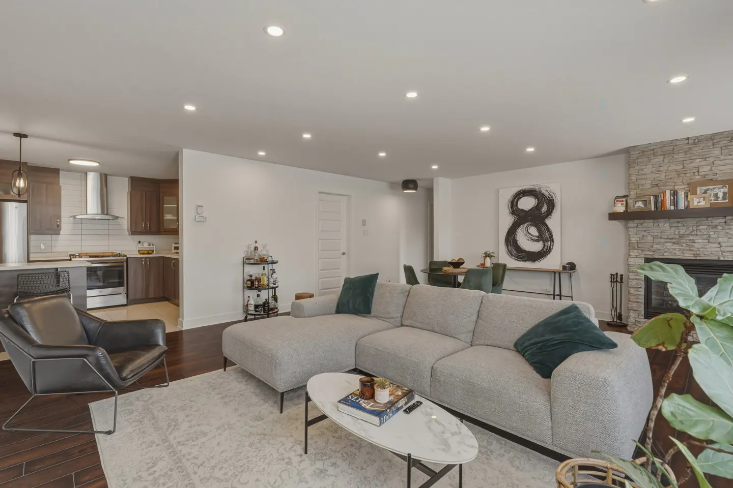 Bright, open-concept living room with a gray sectional sofa, marble coffee table, and a stone fireplace. Kitchen visible in the background.