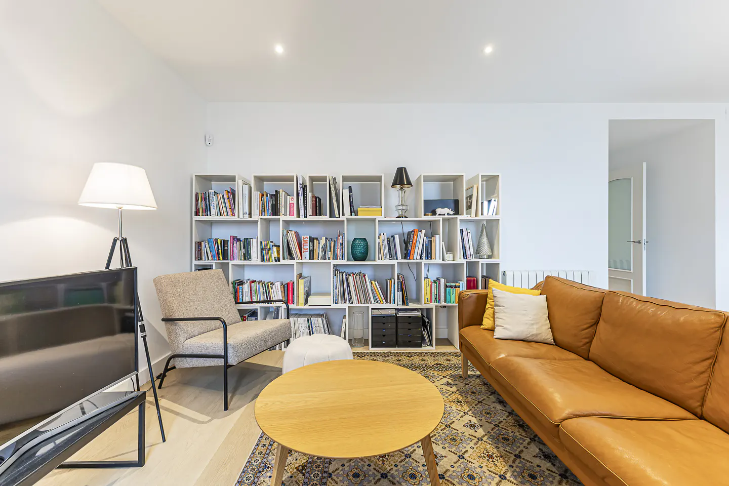 Bright living room with a brown leather sofa, round wood table, and a white bookshelf filled with books.