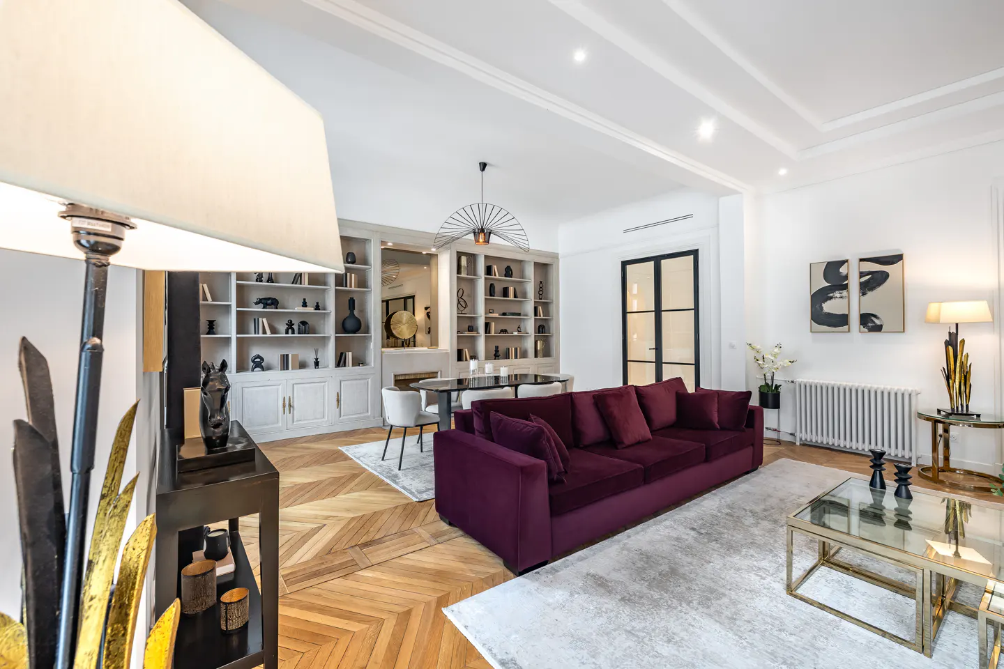 Modern living room with a deep purple velvet sofa, herringbone wood floors, a glass coffee table, and built-in shelving.