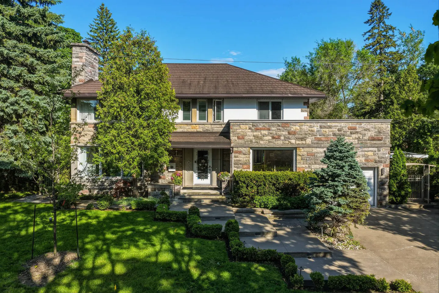 Two-story stone and white house with a brown roof, surrounded by lush green trees, a manicured lawn, and a concrete walkway.