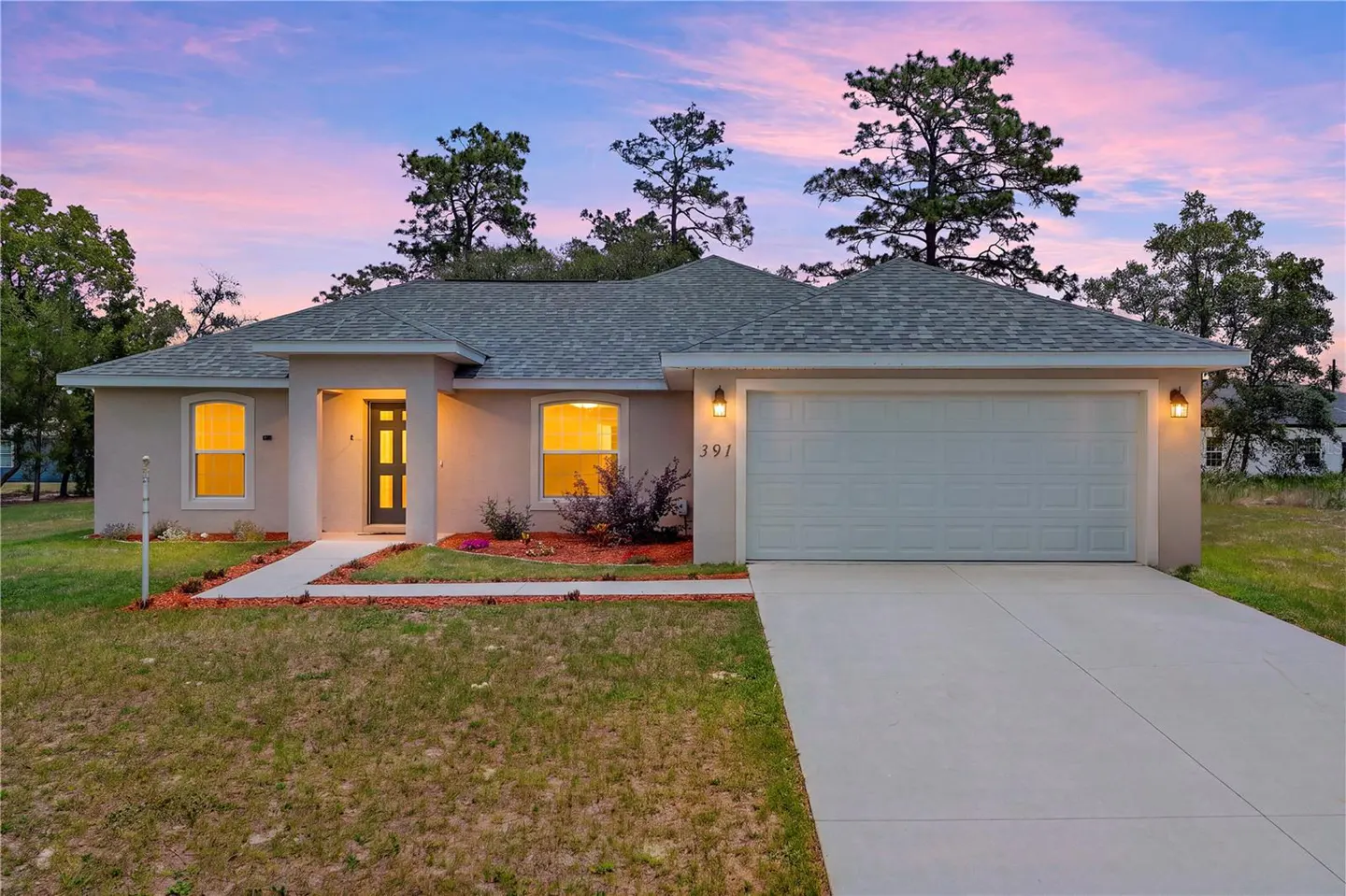 Exterior view of a single-story beige house with a gray roof, white garage door, and a concrete driveway at dusk.