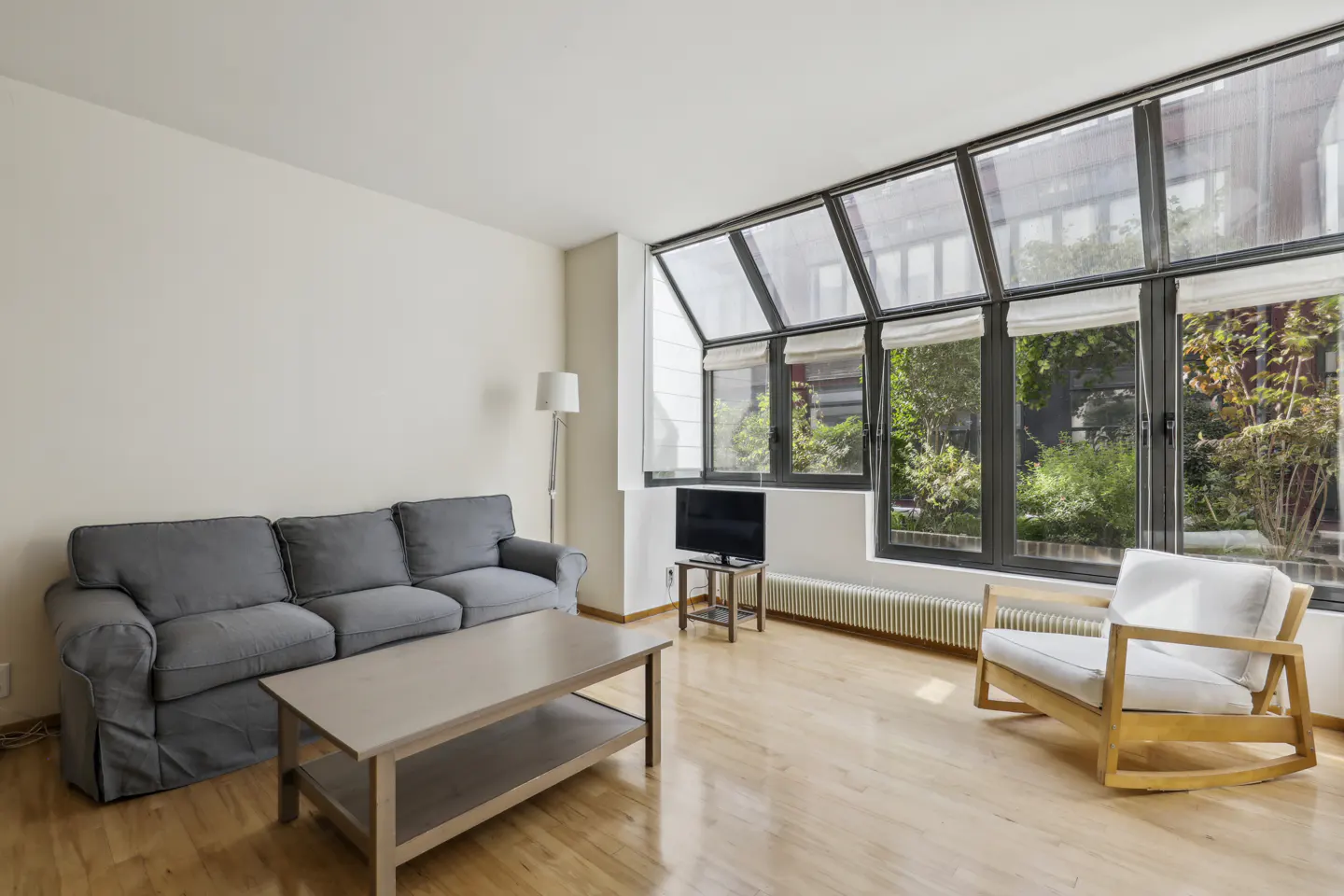 Bright living room with a gray sofa, wood coffee table, and white rocking chair. Large windows overlook a green courtyard.