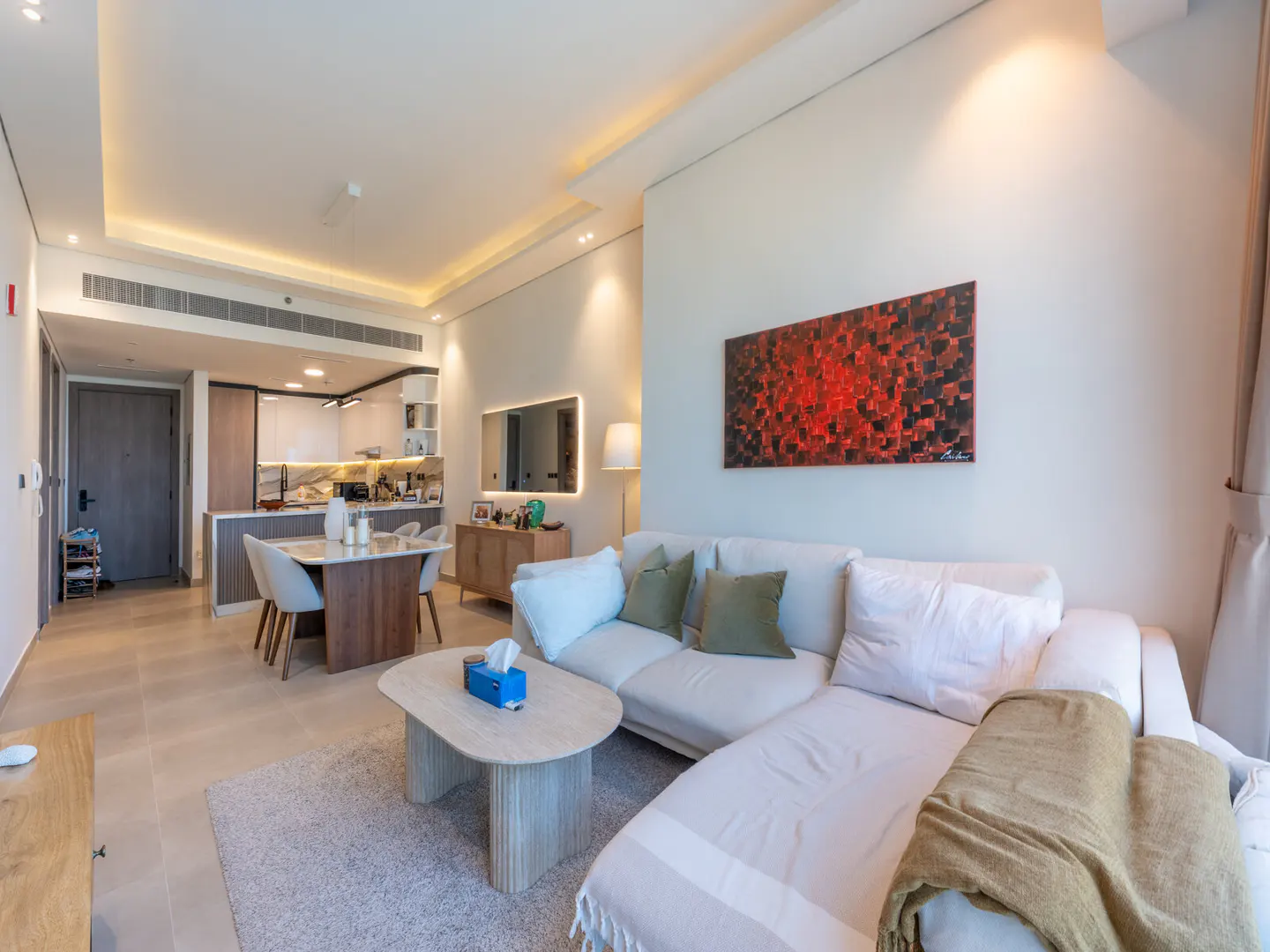 Living room with white sofa, beige rug, and marble coffee table. Red and black abstract art hangs on the wall. Kitchen in the background.