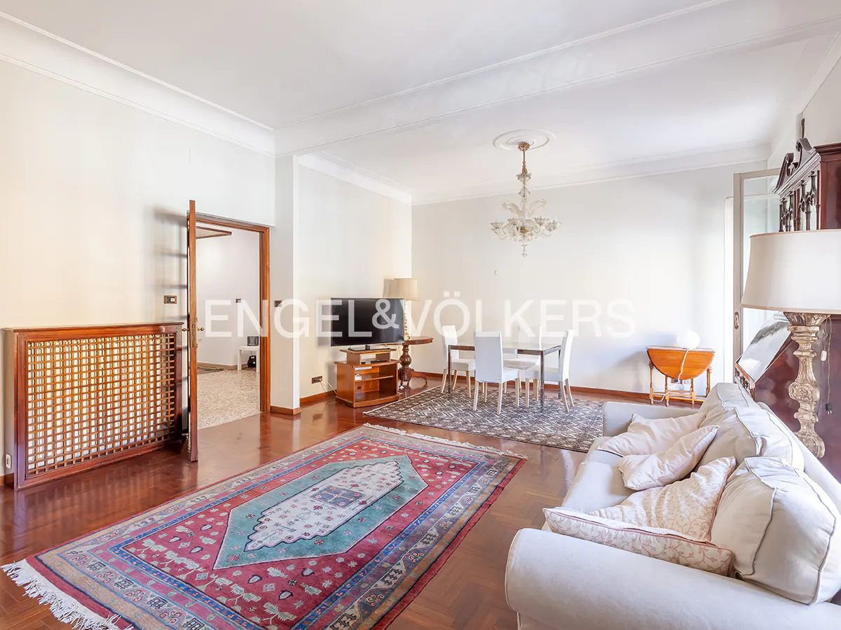 Spacious living room with hardwood floors, a large red patterned rug, white sofa, dining area, and crystal chandelier.