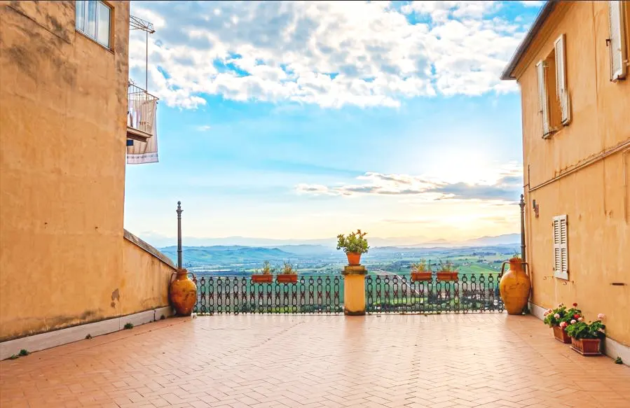 Terracotta-tiled terrace with flower pots and a wrought iron railing overlooking a green valley under a blue, cloudy sky.
