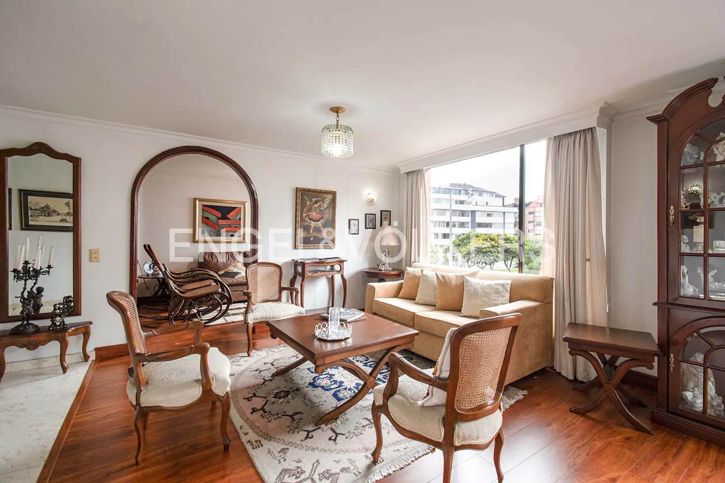 Living room with hardwood floors, beige sofa, chairs, and a square coffee table on a patterned rug. Large window with city views.