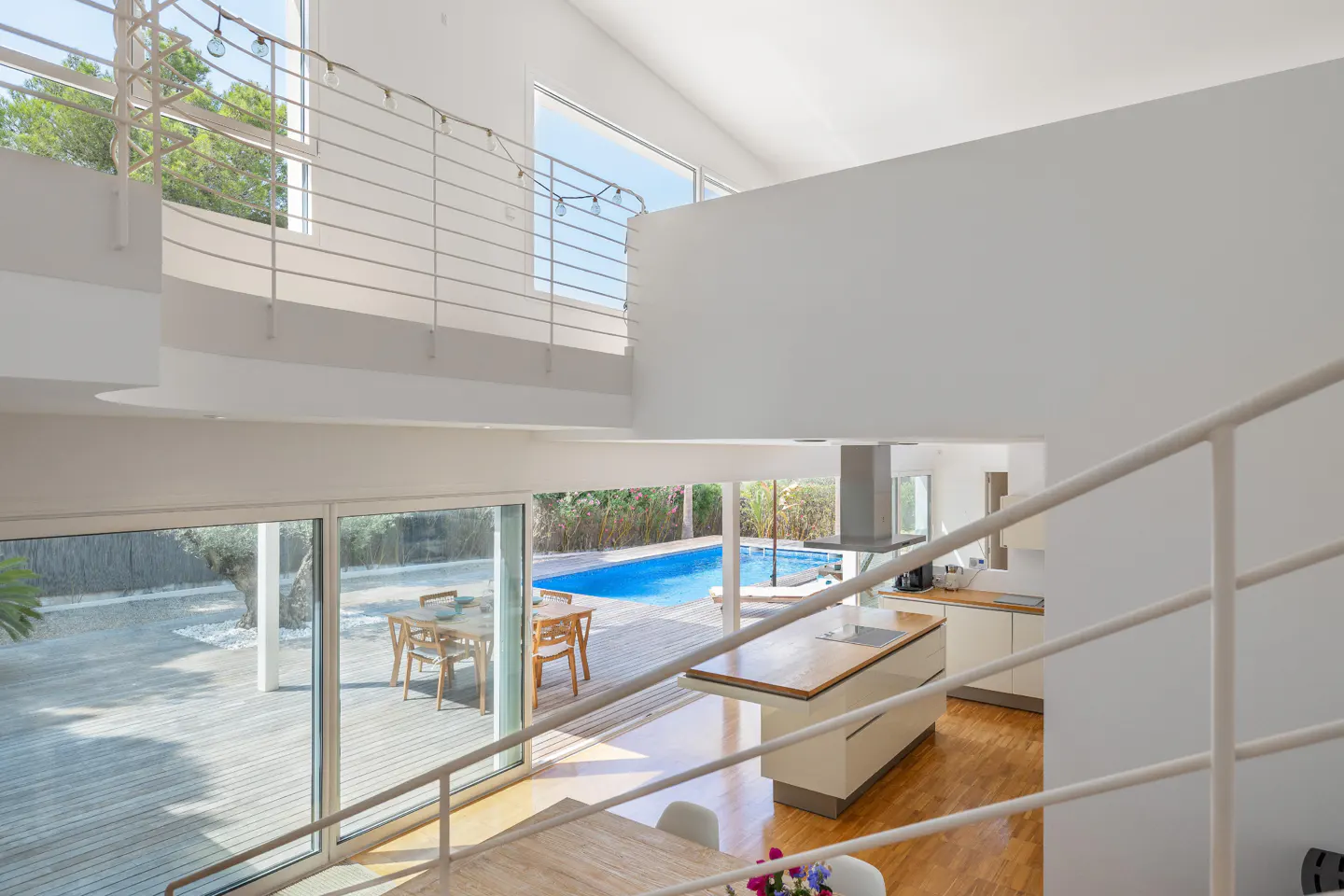 Interior view of a modern white house with a pool visible through large glass doors. A white staircase is in the foreground.