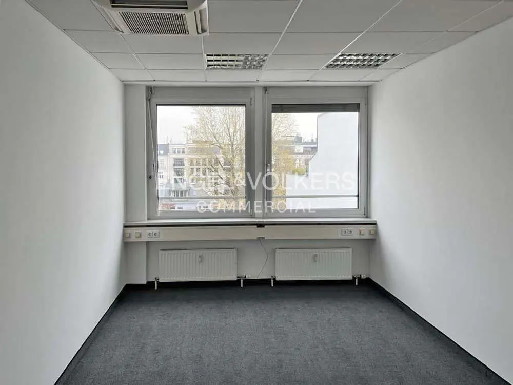 Empty office space with gray carpet, white walls, and two windows overlooking a city. Radiators are under the windows.