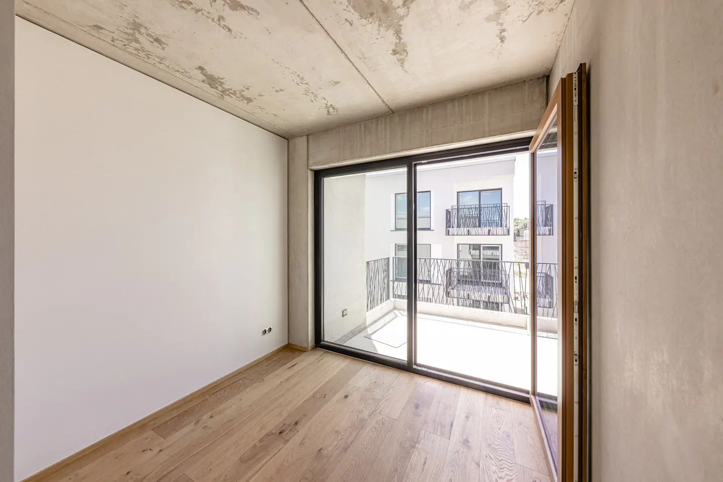 Unfurnished room with light wood floors, white walls, and a concrete ceiling. Sliding glass doors open to a balcony with a view of a white building.