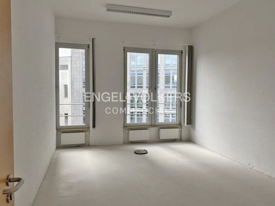 Empty white office space with two windows, radiators, and a light fixture on the ceiling. Engel & Volkers logo is visible.