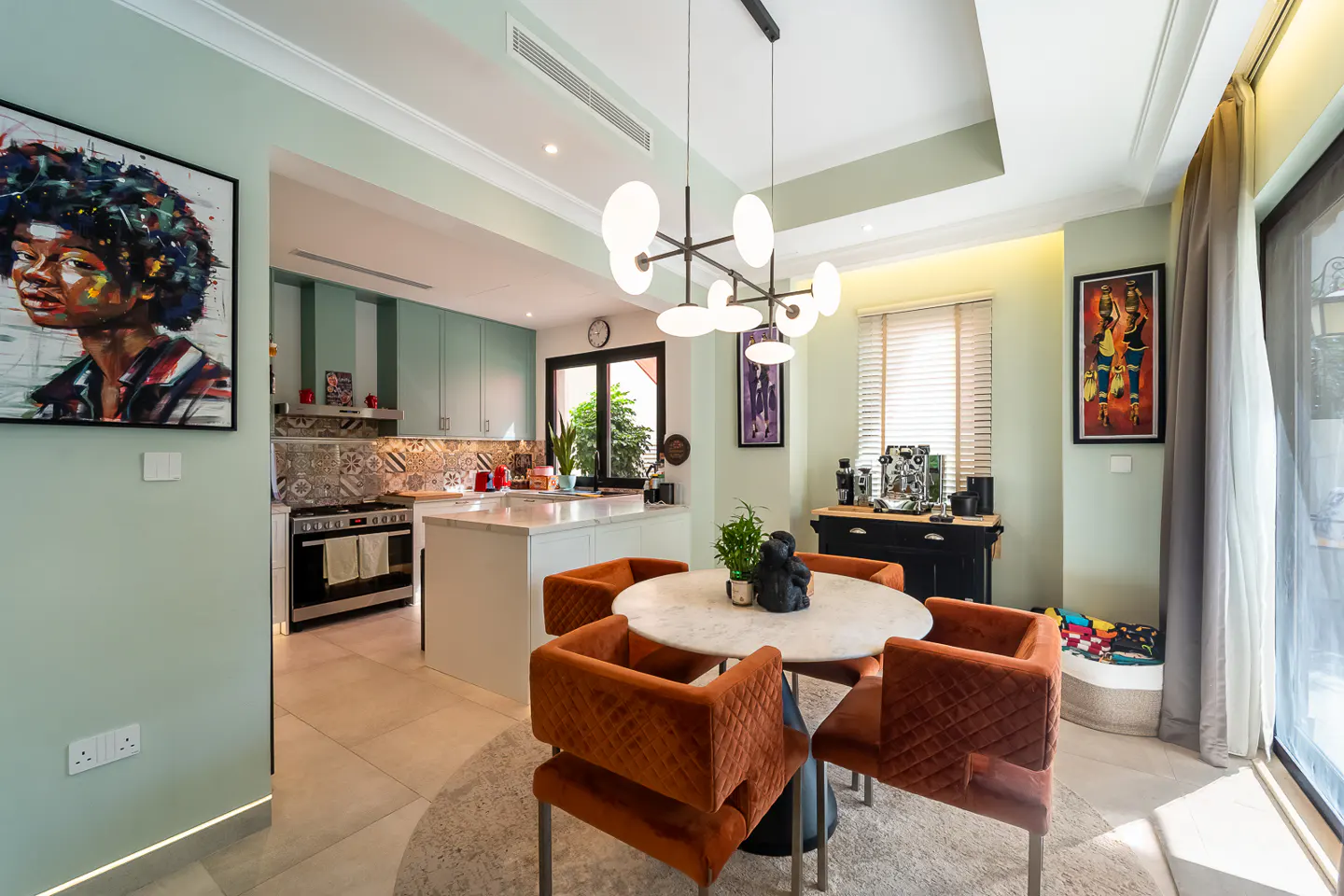 Open-concept kitchen and dining area with mint green walls, modern lighting, and orange velvet chairs around a marble table.