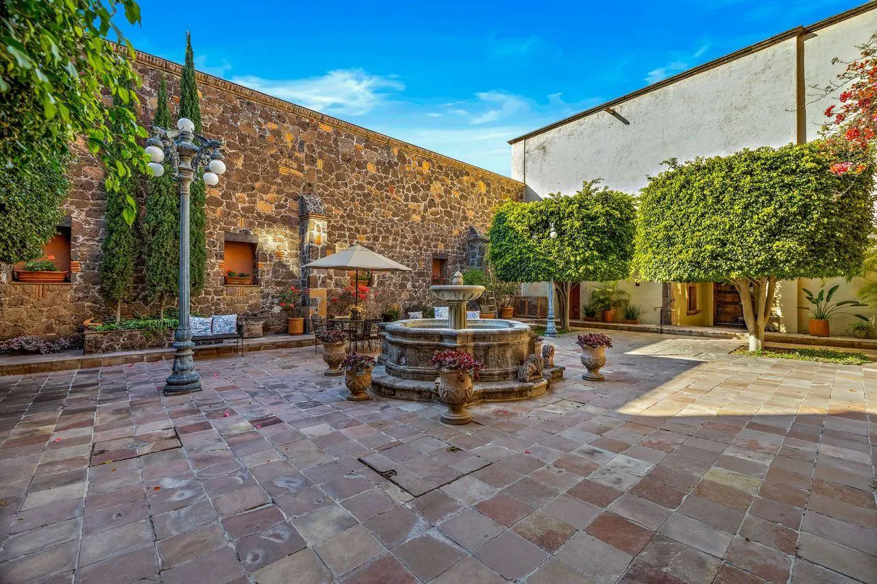 Courtyard with stone fountain, potted plants, and stone walls under a blue sky. A lamp post and trimmed trees add to the ambiance.